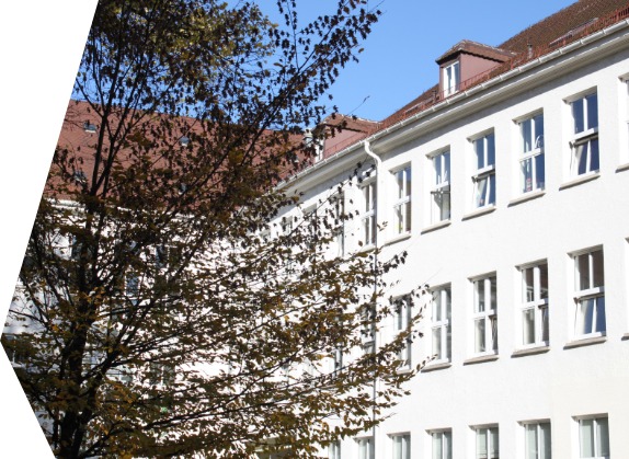 Das Bild zeigt ein weißes mehrstöckiges Gebäude mit roten Dächern und vielen Fenstern. Im Vordergrund ist ein Baum zu sehen, dessen Äste und Blätter einen Teil des Gebäudes verdecken.